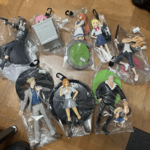 Collection of anime figure figurines in plastic bags laid on a wooden floor, each on stands or bases, in varied poses and outfits.
