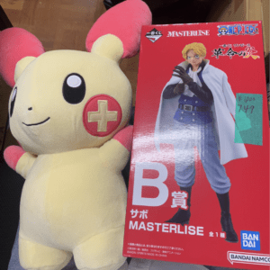 Cream-colored plush with big pink ears beside a red One Piece figure box, showing a blond-clad character in a cape on the cover.