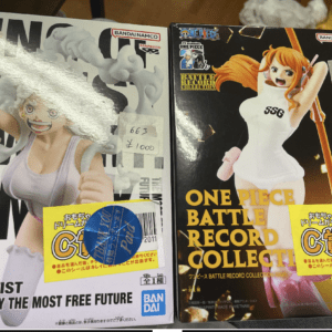 Two One Piece Battle Record Collection figure boxes on a store shelf, side by side; left box shows a pink-clad character, right box shows a white-dress character with orange hair; price tags ¥1000 visible.
