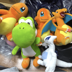 Pile of colorful plush Pokémon toys on a floor, including orange dragon-like figures, a bright green dinosaur, and a white dragon among black bags.
