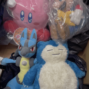 Pile of plush toys in plastic bags: a large pink Kirby, a Mario-like figure, a blue Lucario, and a blue-and-white Snorlax.
