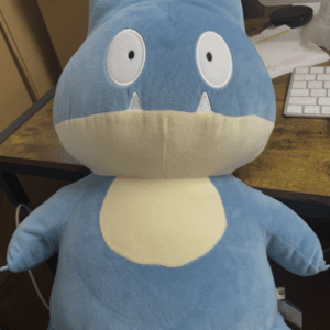 Blue plush dragon toy with a cream belly sitting on a wooden desk near a computer monitor.