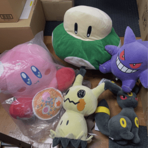 Assorted plush toys piled on the floor beside cardboard boxes, including a pink round character, a green round character, and other whimsical figures.