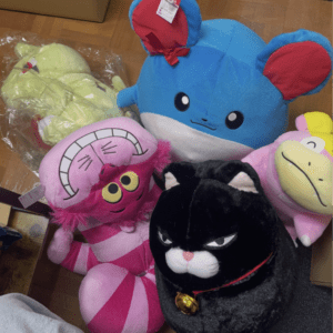Blue plush bear with red inner ears and a bow, surrounded by pink and black stuffed toys on a wooden floor as a pile of plushies ready for play.