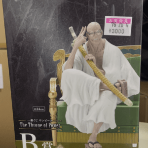 Box for One Piece figure 'The Throne of Power' showing a bald man in a white robe seated on a green throne, gripping a long yellow staff; Bandai Namco logo visible.