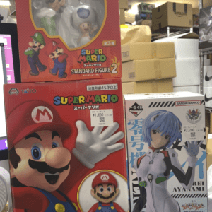 Display of boxed action figures on a store shelf: a large red Super Mario box featuring Mario, with a pink Super Mario Standard Figure 2 box above and another figure box to the right.