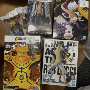 Grouping of anime figure boxes on a table: Naruto Grandista figure (orange suit) at bottom left, One Piece King of Artist The Rob Lucci Awakening Ver. box at bottom right, and two large One Piece Battle Record Collection boxes plus smaller figure packs visible at top, with price stickers on several boxes.