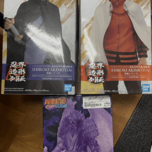 Two boxed Naruto action figures side by side on a wooden surface: left figure in a black cloak holding a sword, right figure Naruto in an orange outfit with a white cape; a purple statue box rests below.