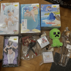 Collection of boxed anime figures and plush on a wooden floor, including SAO E.X.Q. figure and a blue-haired character in a tub box, plus a green Creeper plush.