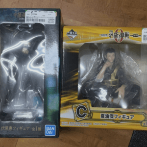Two boxed action figures sit on a wooden floor: one with dark blue packaging labeled 'B' on the left, the other with bright yellow packaging labeled 'C' on the right, both visible through clear windows in the boxes aside from plastic wrap.