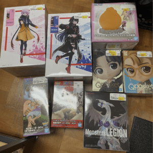 Collection of anime figure boxes arranged on a wooden floor, featuring two large boxed female characters, a yellow-orange figure, and several Qposket and Monsters Legion boxes on display.