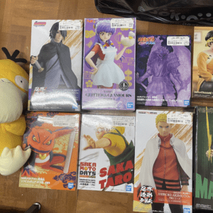 Top-down view of various anime figure boxes laid out on a wooden floor, with a large yellow plush toy on the left.