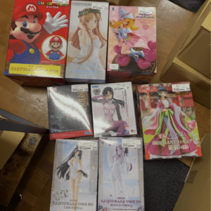 Boxed anime figurines arranged on a wooden floor, featuring a red Mario box and several colorful magical-girl character boxes nearby on cardboard.