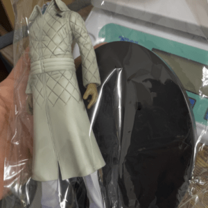 Action figure of a man in a light quilted coat and sunglasses, inside a plastic bag with a black circular stand nearby.