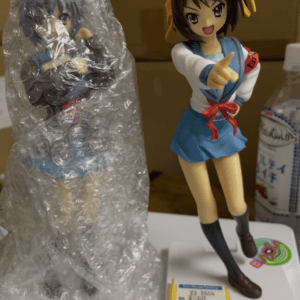 Two anime figures on a shelf: one unwrapped, in a blue sailor uniform pointing forward; the other wrapped in bubble wrap beside it.