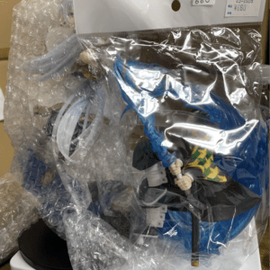 Action figure still wrapped in clear bubble wrap with blue cape visible inside packaging, label showing price in the corner.