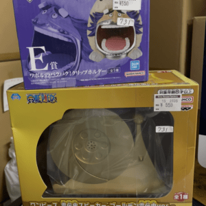 Stacked toy boxes: a purple One Piece 'The Greatest Battle' box with a large open-mouthed creature artwork on top of a yellow speaker-box with a clear window showing a circular speaker inside.