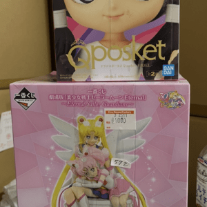 Stacked boxed anime figures: top box shows a large-eyed character from Qposket; bottom box features Sailor Moon Eternal figure in pink packaging.