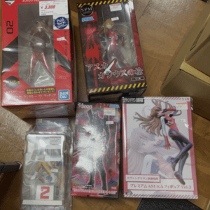 Several boxed anime figures on a wooden floor, including red and pink character boxes and a clear window display on two boxes far left and center-right.
