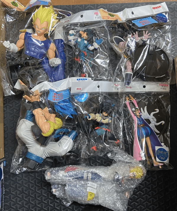 Ver Fotos Recientes 5 - Articulos Japan (post Box Japan) Multiple packaged anime action figures laid out on a dark patterned surface, including a tall yellow-haired character in blue and several other colorful figures. Articulos Japan (Post Box Japan)