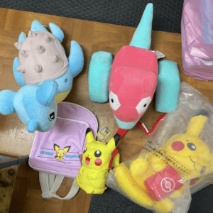 Colorful plush toys scattered on a wooden floor: a blue dragon-like creature with a light gray shell, a pink dragon with turquoise accents, a Pikachu-themed pink bag, and a yellow Charizard-like toy nearby.