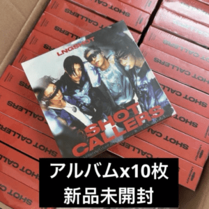 Pile of red boxes stacked in a box, with a glossy album cover showing four young men wearing sunglasses resting on top; Japanese caption overlaid suggests 10 unopened albums.