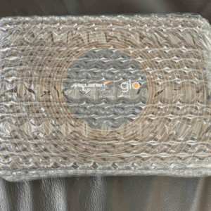 Box wrapped in bubble wrap resting on a dark surface; center shows a circular orange coil pattern with a logo visible through the wrap.