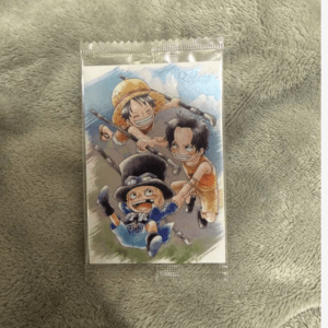 Colorful One Piece card featuring Luffy (straw hat) and two companions in an action pose, sealed in a plastic sleeve on a soft gray blanket.