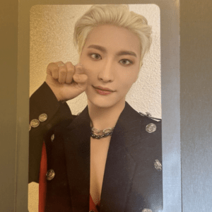 Portrait of a person with bleached hair in a black blazer, posing with a raised fist near the cheek and a subtle smile.