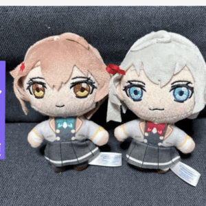 Two anime-style plush dolls in school uniforms sit side by side on a dark gray couch; one has peach hair, the other silver hair, with a purple label on the left.  Articulos Japan (Post Box Japan)