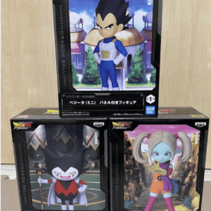 Three boxed Dragon Ball DAIMA figures on a wooden surface: top center features Vegeta in blue armor, with two boxes beneath—one a dark villain with a crown and scepter, and the other a blue-skinned girl with pigtails in an orange outfit.