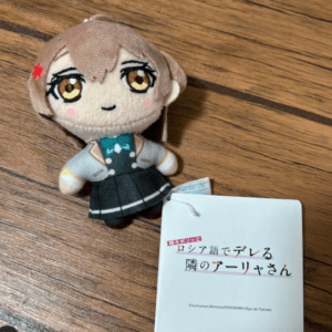 Plush chibi character toy with brown hair in a school uniform on a wooden surface, tag attached to its side (Japanese text).  Articulos Japan (Post Box Japan)