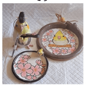 Handmade ceramic plates and a hoop frame with a yellow chick illustration among pink blossoms, displayed on a quilted surface.