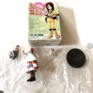 Boxed anime-style figure set with a yellow-clad female character on the front, labeled VOL.5, alongside loose figures in a plastic bag and a black circular stand nearby