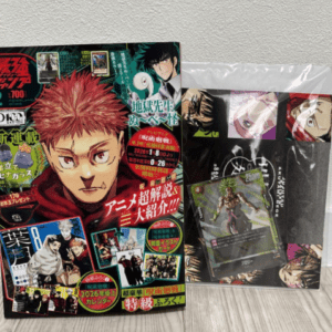Two Japanese manga magazines on a surface, the left cover features a close-up of a red-haired character; a plastic-wrapped bundle of cards sits to the right.
