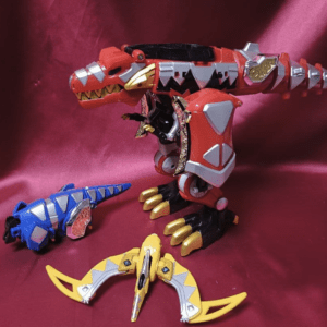 A large red robotic dragon with gray armor plates and gold claws stands on a pink backdrop, with a blue dragon toy and a yellow winged weapon nearby.