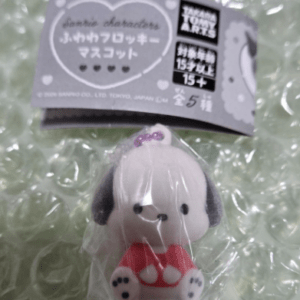 Small plush keychain of a white dog with black ears and a tiny purple bow, wearing a red outfit, in plastic packaging.