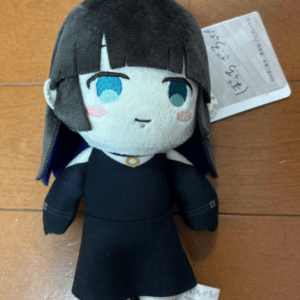 Soft plush doll of a girl with black bob hair, blue eyes, wearing a black dress, on a wooden floor with a tag in the background