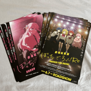 Stack of anime promotional posters with colorful character art; pink-haired girl on the left poster, a group of four characters on the right poster, Japanese text visible, laid on a light surface.