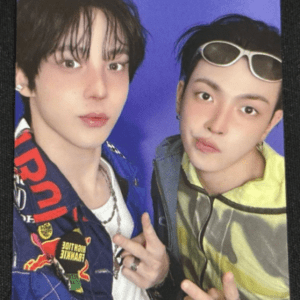 Two young men pose for a photo against a blue background; left wears a patch-covered denim jacket and necklace, right wears a neon jacket with white sunglasses on his head, both making peace/hand gestures.