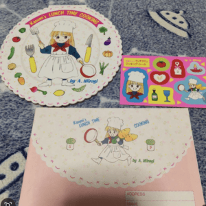 Illustration of a girl chef on a scalloped plate with vegetables and utensils, titled 'Kasumi's Lunch Time Cooking' and a recipe sticker sheet nearby.