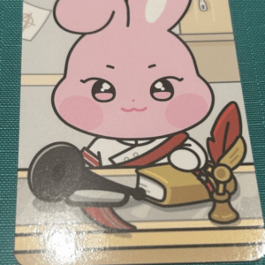 Cute pink bunny in a white outfit with a red sash sits at a desk, touching a gold ceremonial object with a red feather in front of it.