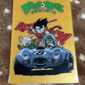Dragon Ball trading card in a clear sleeve with a yellow background; shows Goku in a blue vintage race car, flanked by a mouse-like character and a helmeted rider, racing forward.