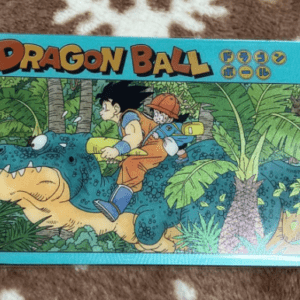 Dragon Ball branded card showing Goku and a friend riding a blue dinosaur through a jungle, with palm trees and jungle foliage in the background.