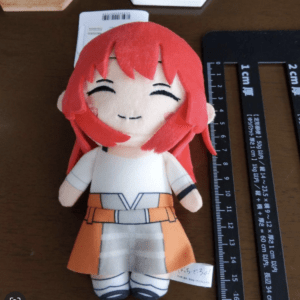 Smiling plush doll with red hair, white top and orange skirt, placed on a wooden table beside a ruler  Articulos Japan (Post Box Japan)