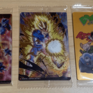 Three Dragon Ball trading cards in plastic sleeves: left shows a blue armored character with energy blasts; center features a character with a bright yellow aura; right shows Goku in a car on an orange card