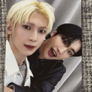Close-up selfie of two people smiling, one with bleached blond hair in a light jacket and the other with dark hair in a black outfit, leaning in together.