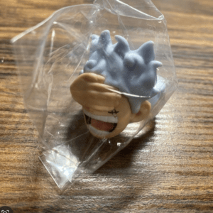 Small cartoon-style figure head with blue spiky hair, wrapped in clear plastic, on a wooden surface.