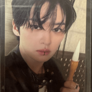 Portrait of a person with dark hair and makeup, wearing a black leather jacket, holding a white dagger-like prop near their face.