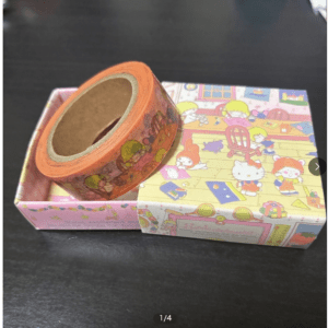 Orange decorative tape roll resting on a pink Hello Kitty–themed gift box with colorful cartoon illustrations.